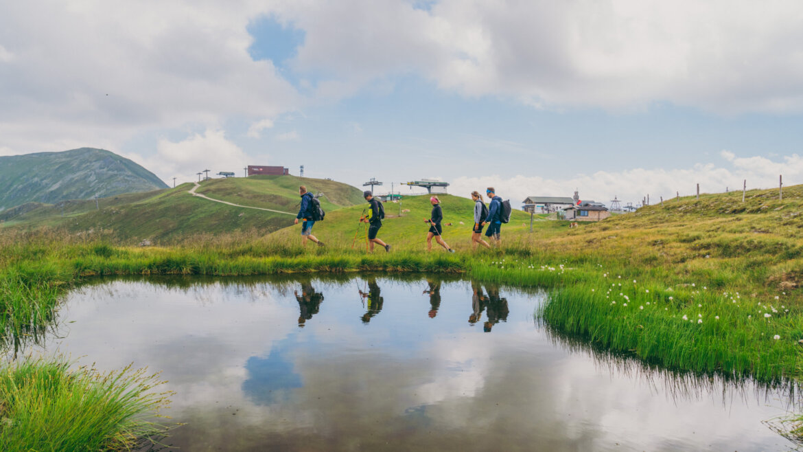 (c) Peter Maier Four walkers walk beside a small pond in a grassy mountain landscape under a partly cloudy sky. (Enlarged view)