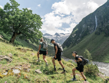 Three people hike up a grassy hill in a mountain valley with trees and a distant waterfall under a cloudy sky.