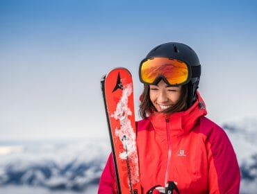 Woman in red ski jacket and goggles holding skis, smiling with snowy mountains in the background.