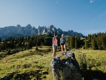 Two women stand on a rock in a grassy mountain landscape with pine trees and jagged peaks in the background.