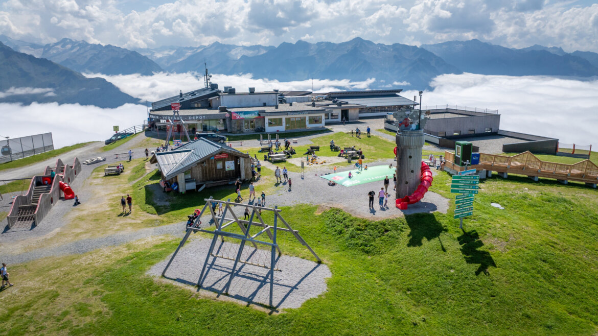 (c) Wildkogel Arena Hilltop playground and buildings with people, surrounded by mountains and clouds below under a bright sky. (Enlarged view)