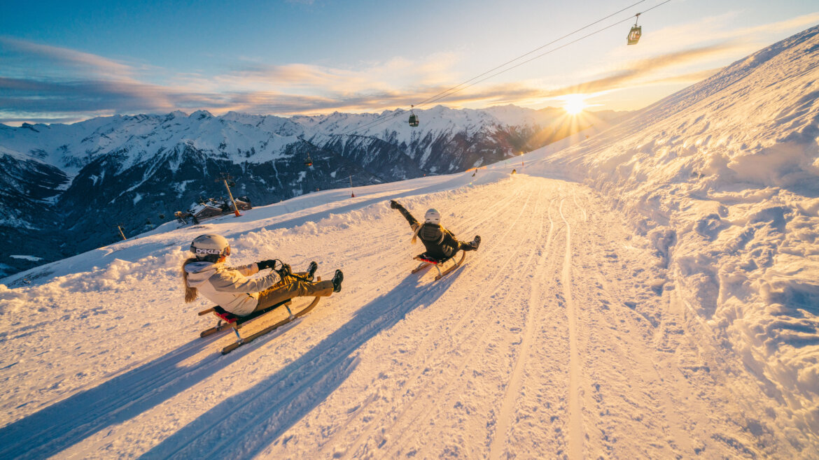 (c) Peter Maier Two people sledging downhill on a snowy mountain at sunset, with mountains and ski lifts in the background. (Enlarged view)
