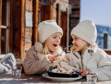 Two young children in winter clothes share a meal outdoors, smiling and savouring the taste of winter.