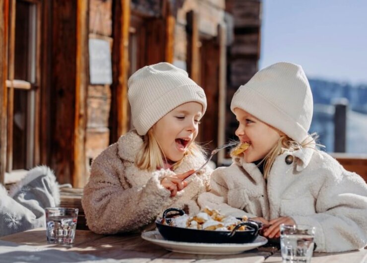 Two young children in winter clothes share a meal outdoors, smiling and savouring the taste of winter.