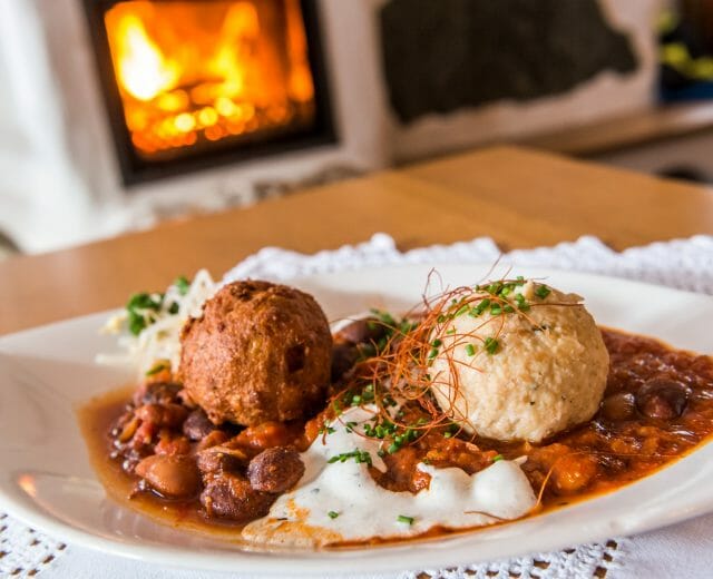 Chili con Carne mit Knödel A plate of beans with two dumplings, garnished with herbs, in front of a cosy fireplace.