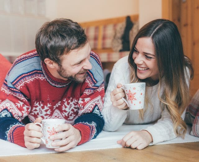 Pärchen mit Heißgetränk A man and woman lying on the floor, smiling and holding festive mugs, enjoying a cosy moment together.
