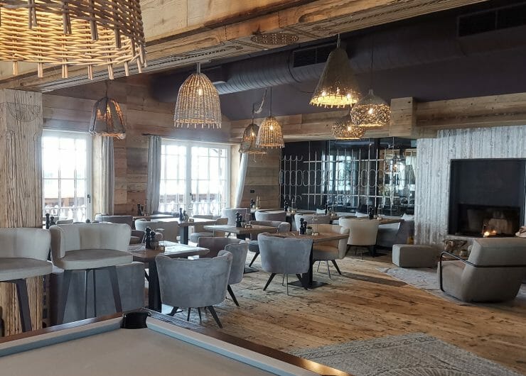 Cosy lounge with wooden walls, modern chairs, wicker lights, and a fireplace, lit by natural daylight.