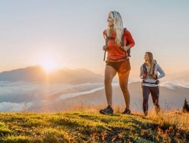 Two women hiking on a grassy hilltop at sunrise with mountains and clouds in the background.