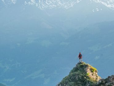 A hiker stands on a mountain peak, overlooking a valley with distant snow-capped mountains.