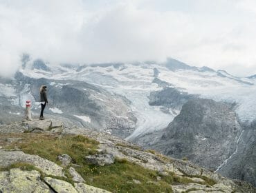 Person standing on rocky hilltop, overlooking snowy mountains and glacier under a cloudy sky.