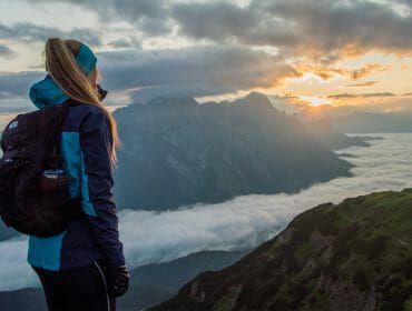 saalfelden-leogang-fb (12 von 17) Woman in hiking gear stands on a mountain, overlooking a cloudy valley at sunrise or sunset.