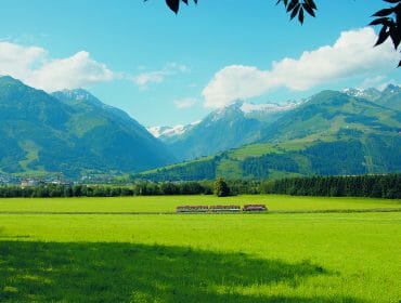 A red train passes through a green field with mountains and forests in the background under a blue sky.