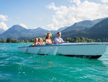 A family of four boating on a turquoise lake with mountains and trees in the background under a blue sky.