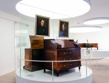 A wooden antique desk and two framed portraits displayed in a modern, glass-enclosed museum exhibition.