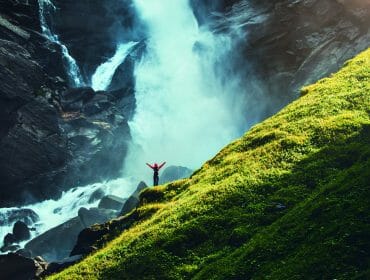 Person with arms raised stands near a large waterfall, beside a steep, grassy hillside.