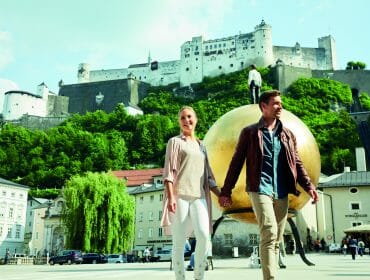 A couple hold hands and walk in front of a golden sphere with a fortress on a hill behind them.