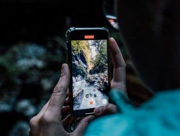 A person records a scenic forest stream on their phone whilst standing outdoors.