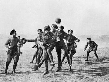 stille-nacht-Martin-Reiter-Weihnachtsfriede-Front Soldiers in uniform play a game of football on a pitch, jumping and reaching for the ball in mid-air.