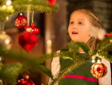 A young girl admiring a decorated Christmas tree with red baubles and lights.