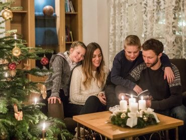 Familie an heilig Abend A family of four sits by a lit Christmas tree and candles, reading together in a cosy living room.