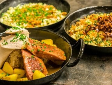Three pans with sausages, roast potatoes, sauerkraut, and two types of cheesy pasta with crispy toppings.