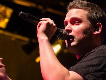 A man sings into a microphone on stage under warm lighting, wearing a black shirt.