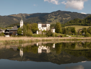 The village Goldegg with a church, castle, and trees reflected in the calm lake "Goldeggersee", mountains in the background.