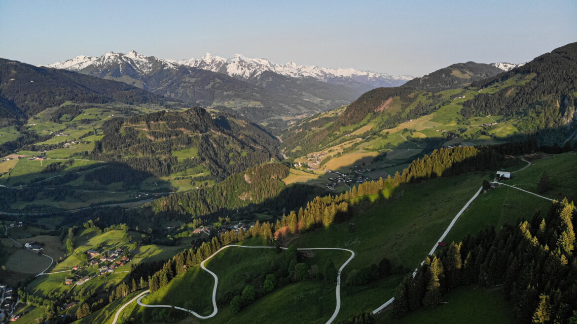 A winding road cuts through green hills along the Salzburger Sonnenterrasse with mountains and snowy peaks in the background under a clear sky.