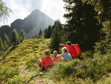 Four people in hiking gear sit on a grassy slope, looking at a mountain peak surrounded by trees.