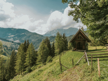 A small wooden chapel stands on a grassy hillside with mountains and pine trees in the background.