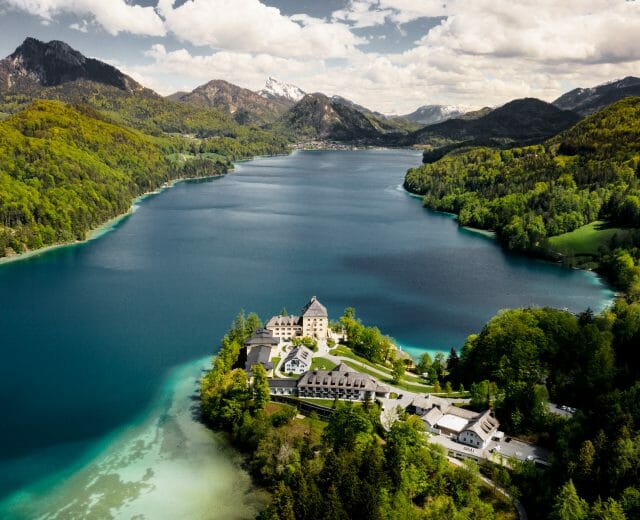 Aerial view of a lake surrounded by mountains, with a castle-like building on a forested peninsula.
