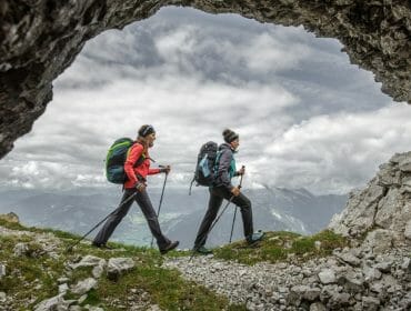 Two hikers with rucksacks and walking sticks trek through a rocky mountain cave, with clouds and peaks in the background.