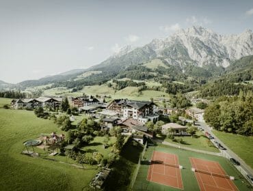 Aerial view of a mountain village with tennis courts, playground, green fields, and towering mountains in the background.