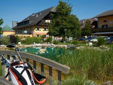 Golf bag on a wooden bridge overlooking a pond, with buildings and parked cars in the background on a sunny day.