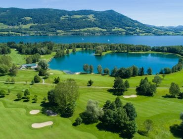 Aerial view of a golf course with bunkers, trees, a pond, and a lake surrounded by hills.