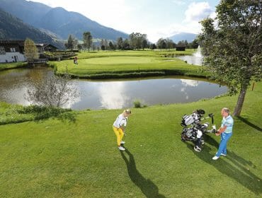 Two people playing golf near a pond, with mountains and trees in the background on a sunny day.
