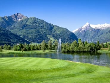 GC Zell_am_See_-15 Golf course green beside a lake with fountain, surrounded by mountains and trees under a clear blue sky.