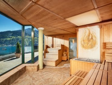 Grandhotel Zell am SeeFinnische_Sauna_ohne_Menschen Wooden sauna room with large windows overlooking a lake, mountains, and a sunny outdoor terrace with potted plants.