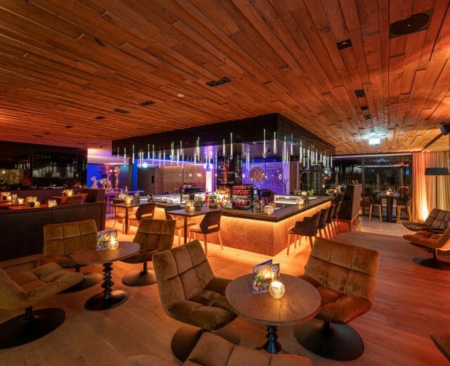 Modern lounge bar with warm lighting, wooden decor, cosy chairs, and tables set for guests.