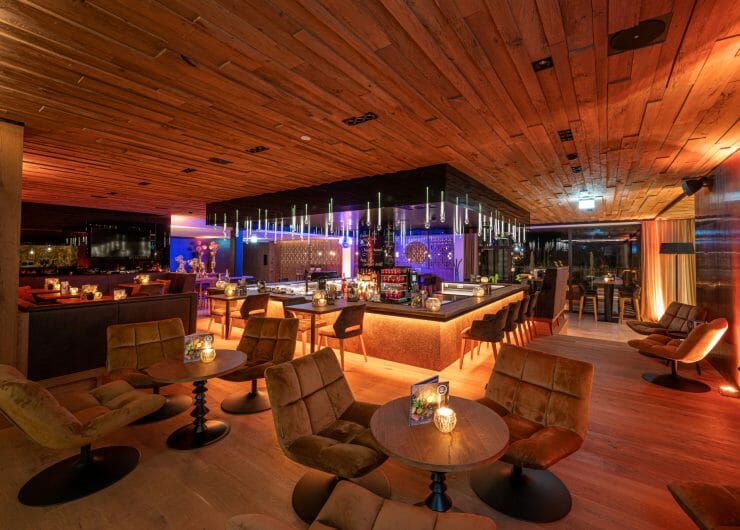 Modern lounge bar with warm lighting, wooden decor, cosy chairs, and tables set for guests.