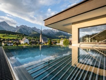 Infinity pool overlooking an alpine village, with mountains and a church spire in the background.