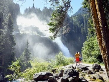 A family stands on a forest trail, admiring a large waterfall surrounded by trees and mist.