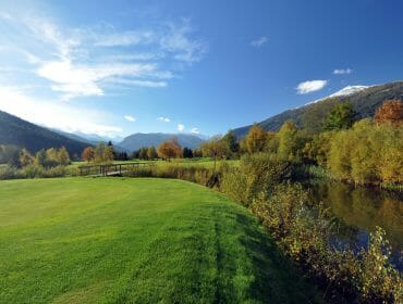 Green golf course with a small wooden bridge, a pond, trees, and distant mountains under a blue sky.
