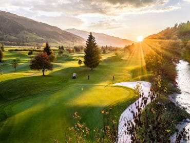 A scenic golf course at sunset with mountains, trees, a river, and two people near a golf buggy.