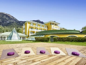 Four yoga mats with purple cushions on a wooden decking outside a yellow building with mountains in the background.