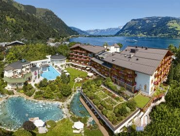 A lakeside hotel with gardens, a swimming pool, and mountain views under a clear blue sky.