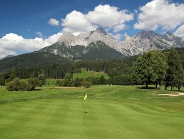 copyright_ales_fevzer-1170×658 Golf course with a flag on the green, trees, and mountains in the background under a blue sky with clouds.