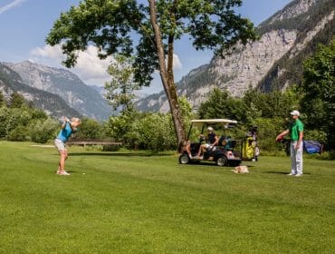 Person swings golf club on a green course as two others watch by a golf buggy; mountains in background.