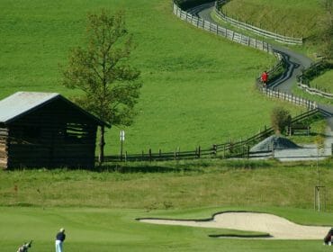 Two golfers on a green field near a sand bunker, with a wooden shed and winding road in the background.