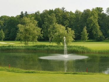 A golf course with a small pond, a fountain in the centre, and a yellow flag in the foreground.